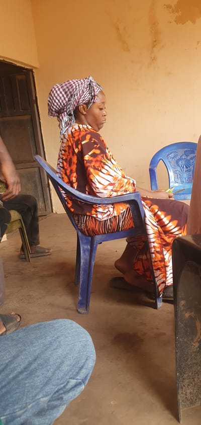 The image captures a candid moment, likely during the daytime, in Idah, Nigeria. The central subject is an adult woman, appearing to be of African descent, seated comfortably in a blue plastic chair. She is dressed in a vibrant, flowing garment featuring an orange, brown, and white abstract pattern, possibly resembling animal stripes. Her head is adorned with a checkered headwrap in red, white, and dark blue/black plaid. Her head is tilted back, resting against the chair, and her eyes appear to be closed or looking upwards, conveying a sense of rest or contemplation.
The setting appears to be an outdoor or semi-outdoor space, characterized by a plain, light brown or tan wall in the background, showing some minor smudges or discoloration. The ground beneath is a similar earthy brown, suggesting dirt or compacted soil. Another blue plastic chair with an ornate backrest is partially visible behind the woman, and a dark, heavy object, possibly a table or furniture, is seen in the upper right.
The presence of other individuals is indicated by partial views of their limbs: a leg clad in blue jeans is visible on the right, and another person's legs in dark trousers with sandals, along with their hands, are seen in the lower left portion of the frame. This suggests a small gathering or social interaction. A dark door or gate is also partially visible towards the bottom left. The overall atmosphere is informal and relaxed. No discernible text is visible in the image.