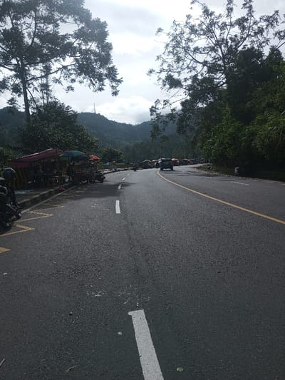 The image displays a two-lane asphalt road traversing a mountainous landscape in Sandamal Eliya, Sri Lanka. The sky is overcast with visible clouds, and lush green, densely forested mountains dominate the background, featuring a communication tower on the peak of the furthest mountain. The road is marked with a continuous yellow line separating the lanes and broken white lines on the left shoulder. Along the left side of the road, behind a yellow and black striped curb, a series of vendor stalls are set up. These stalls are covered by assorted tarpaulin awnings, including red, green, and multi-colored designs, displaying various goods. Two motorcycles are parked on the far left, one with a helmet resting on its seat. On the road, a dark SUV is seen moving away from the viewer in the right lane, and additional vehicles, including motorcycles, are visible further in the distance.