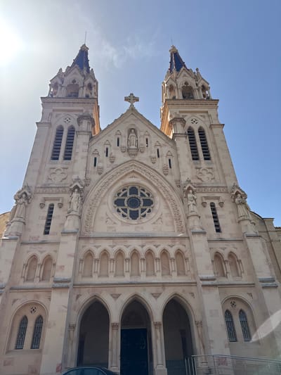 This is a low-angle, wide shot of the exterior of a large, ornate church building in Benirredrà, Spain, under a bright, clear blue sky. The church is constructed of light-colored stone and features a prominent, symmetrical facade with two tall, Gothic-style bell towers flanking a central structure. Each tower has a dark, pointed roof. The central facade is adorned with a large rose window above a series of arched entrances. Statues of figures in religious attire are visible in niches on the facade and on the upper levels of the towers. The architecture displays intricate detailing, including pointed arches, decorative carvings, and small, narrow windows. Sunlight casts bright highlights on the building and creates some glare in the upper left corner of the image. A sliver of a car is visible in the lower foreground.