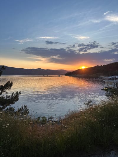 The image depicts a sunset over a large body of water, identified as being in the city of Brosh, Bulgaria. The sun, a bright orange-yellow orb, is positioned low on the horizon, partially obscured by a dark, forested hill on the right side of the frame. Its intense light casts a strong glare and reflection across the water, creating a vibrant orange and yellow hue that fades into cooler blues and greys further from the sun's path.
The sky above transitions from deep orange and yellow near the horizon to a lighter blue and white with scattered cirrus and cumuliform clouds higher up. Some clouds are dark grey, particularly in the middle and upper-left sections.
In the mid-ground, a range of dark, silhouetted mountains or hills stretches across the far bank of the water. Several small, white, rectangular structures are visible floating on the water's surface in this area, likely aquaculture facilities or floating docks.
On the immediate right side, a white, multi-level structure with windows, resembling a house or lodge built over the water on docks, is prominent. Yellow kayaks or small boats are tethered next to it. The shoreline in the foreground, particularly on the right, is covered with dense, tall grasses and numerous small white wildflowers. Dark branches and needles of a coniferous tree partially frame the lower-left corner of the image. The water surface exhibits gentle ripples, reflecting the sky's colors and the ambient light. No people are visible.