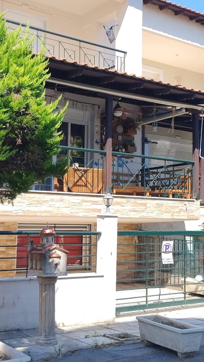 The image captures a sunlit scene in Neoi Epivates, Greece, focusing on the exterior of a building. In the foreground, a decorative concrete column supports a miniature model of a church with a copper-colored roof. Behind this, a railing with green metal bars runs horizontally, separating the ground level from the upper floor. A window with a red-tinted glass is visible on the left.

The building's facade is adorned with light-colored, textured stone blocks. Above this, a balcony features a dark-colored railing with a diamond pattern and a wooden overhang supported by dark posts. The balcony itself contains outdoor furniture, including a wooden table and benches. A large green conifer tree partially obscures the left side of the building.

Further back, a green metal fence encloses an area, and a circular sign with a red 'R' inside, along with Greek text indicating "NO PARKING," is attached to it. A concrete planter box sits on the ground in front of the fence. The sky above is clear and blue.