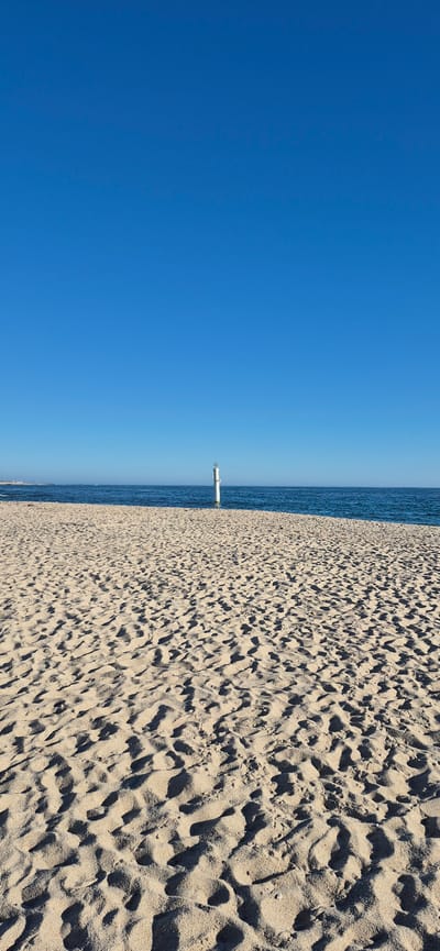 A wide-angle, eye-level shot captures a serene beach scene under a clear, bright blue sky. The foreground is dominated by textured sand, showing a pattern of ripples and small dunes created by the wind, casting subtle shadows. In the mid-ground, the calm, dark blue ocean meets the sandy shore, with a thin line of distant land visible on the far left. A solitary white pole with a light fixture stands in the water, positioned off-center to the right. The overall atmosphere is one of peacefulness and natural beauty. This location is the beach in Vila do Conde, Portugal.