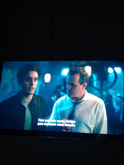 This media file is a photograph taken in El Espinal, Venezuela, capturing a screen displaying a scene from a movie or TV show. The screen shows two men in an indoor setting, likely a dimly lit workshop or industrial space, characterized by blurred blue-tinged lighting and machinery in the background.
On the left is a younger man with dark, curly hair and light eyes, wearing a dark jacket over a lighter shirt. He looks slightly to his right, with a thoughtful or slightly concerned expression. On the right is an older man with lighter, shorter hair, wearing a white collared shirt and a dark tie. He is looking towards the younger man, his expression appearing serious or contemplative.
The lighting in the scene is predominantly blue or teal, creating a dramatic and atmospheric mood. Several bright, diffused light sources are visible in the blurred background.
At the bottom of the screen, white Spanish subtitles are visible, reading: "Pero te tomas mucho tiempo para descubrir cómo hacerlo." This translates to "But you take a long time to figure out how to do it." The photograph itself captures the screen with some surrounding darkness, suggesting it was taken in a dark room.