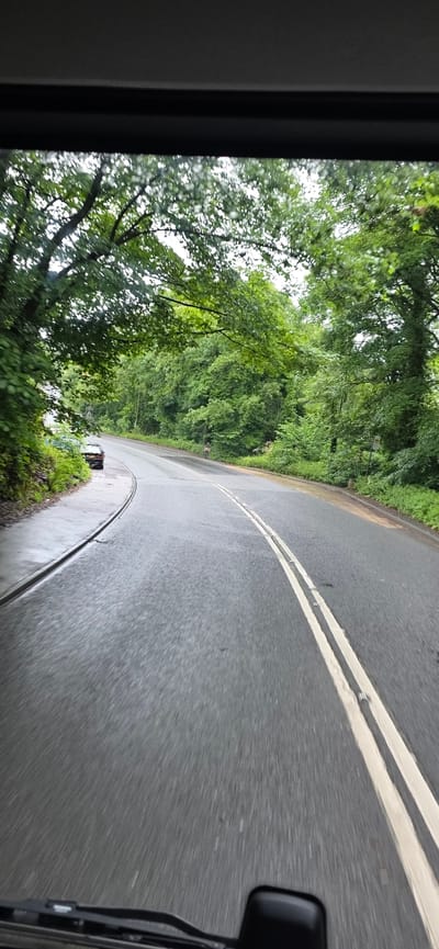 A perspective shot from inside a vehicle, looking out at a wet, winding road flanked by dense green foliage. The road has double white lines painted down its center, and the asphalt appears dark and slick, suggesting recent rain. On the left side of the road, there is a parked dark-colored car and a retaining wall covered in greenery. Overhanging trees create a canopy over the road in the foreground, with their leaves blurred due to motion. The sky is overcast, contributing to the subdued lighting. There are no people visible in the frame. The location is identified as Tameside, United Kingdom.