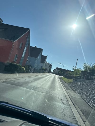 The image shows a street scene in Weidenhausen, Germany, taken from a low angle, likely from inside a vehicle. The sun is shining brightly, casting long shadows and creating lens flare across the sky, indicating it is likely daytime. The sky is a clear blue with a few faint contrails.
On the left side of the street, several houses are visible. The closest house is red with a dark gray roof and white-framed windows. To its right, a blue house with a dark roof and white-framed windows is partially visible. Further down the street, more houses and trees can be seen. The houses appear to be typical residential buildings.
The street itself is paved with asphalt and shows some signs of wear, with cracks and unevenness in places. A concrete curb separates the road from a gravel area or embankment on the right side of the frame. A metal railing runs along the top of this embankment. A street lamp is also visible in the distance. The scene conveys a sense of a quiet residential area on a sunny day. There is no visible text in the image.