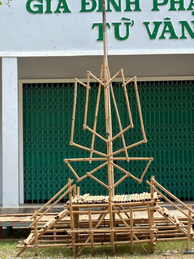 An outdoor scene features an intricate bamboo structure standing prominently in the foreground, likely an artistic installation or temporary decorative piece. The structure consists of numerous bamboo poles joined together with twine or wire. A central vertical pole supports radiating arms that form a large, wheel-like or star-shaped element, creating a complex geometric pattern. To the right, another part of the bamboo construction appears more scaffold-like, with horizontal and vertical poles forming a stable base.
In the background, a large green metal roller shutter door provides a textured backdrop, indicating a commercial or institutional building. To the far left, a portion of a white building is visible, featuring large, raised green letters. The text, in Vietnamese, reads "GIA ĐÌNH PHẬT TỬ VĂN THÀNH," which translates to "Văn Thành Buddhist Family" or "Văn Thành Buddhist Youth Group," suggesting a religious or community center. Patches of green grass are visible on the far right, indicating the ground surface.
The image is captured during the daytime under bright, clear conditions, as evidenced by the sharp details and lack of harsh shadows. No people are visible, focusing attention solely on the constructed environment. The detailed craftsmanship of the bamboo structure, set against the green shutter and white building, implies a cultural or ceremonial context, aligning with the location context of Cam Ranh, Vietnam.