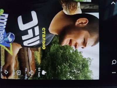 The image shows a screen displaying a video featuring a man wearing a black UFC t-shirt. The background appears to be a green outdoor area with trees. On the screen are also user interface elements like like counts (“54 тыс.”), comments (“Не...тся,” “508”), share (“Под...ься”) and text in Russian (“сталью,” “ПОПОЛНЯЙ”). Based on the appearance of Cyrillic letters, the context of the image may be related to a Russian-speaking audience. The image is a close-up shot.