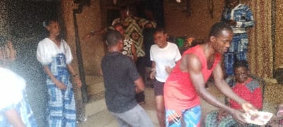A group of people gathers inside or near a building. Some stand or dance in the background, including one individual in a patterned green and yellow top. In the foreground, a man in a red tank top holds a platter of food, appearing to serve it. Another woman sits nearby in a red and gray top. A young woman in a blue and white dress stands off to the side. The scene has a casual, perhaps festive, atmosphere.