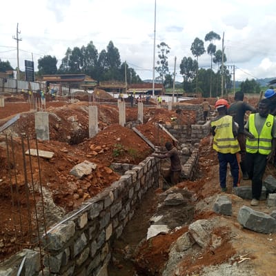 The image depicts a construction site in Borabu Chitago ward, Kenya. Several construction workers are present, some wearing high-visibility vests and hard hats. One worker is laying stones to build a wall, while others appear to be moving materials or supervising the work. The construction site has exposed red soil, partially constructed stone walls, concrete pillars with exposed rebar, and piles of building materials. The surrounding environment includes trees, power lines, and buildings, suggesting the site is located within a populated area.