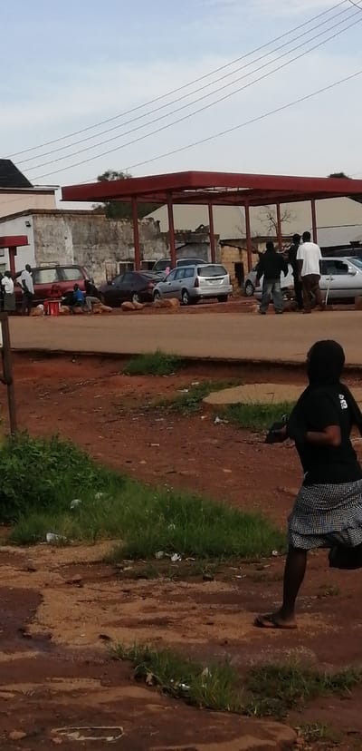 The image depicts a scene at the Kassa Market in Nigeria, characterized by a dusty, reddish-brown dirt road and sparse vegetation. In the foreground, a young person in a black t-shirt and a checkered skirt runs away from the camera. Their posture suggests they might be engaged in some activity or reacting to something.

Further back, in the mid-ground, several individuals are gathered near a red-roofed structure, possibly a gas station or a market stall. Multiple vehicles are parked in the vicinity, including a maroon sedan, a silver hatchback, and a larger maroon SUV. Some people are standing near the vehicles, while others appear to be seated. One individual, wearing a white shirt, is standing near the maroon SUV, and another person in a light blue shirt is seated on the ground.

The background reveals buildings with rough, textured walls and a cloudy sky with visible power lines stretching across. The overall atmosphere suggests a busy, everyday scene within the market environment, with people and vehicles interacting in the open space. The ground is uneven and appears to be a mix of dirt and small stones, common in such outdoor market settings in Nigeria.