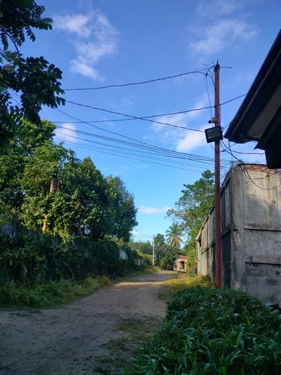 The image depicts a dirt road winding through a rural or suburban area, likely in Kidapawan, Philippines, under a bright blue sky with scattered white clouds. Lush green foliage lines both sides of the road, with trees and plants creating a verdant canopy in some areas. To the right, a concrete wall of a building is visible, adjacent to a weathered red pole supporting electrical wires, a security camera, and a floodlight. Further down the road, a small yellow vehicle, possibly a tricycle or jeepney, is parked. The overall scene suggests a peaceful, sunlit day in a tropical setting.