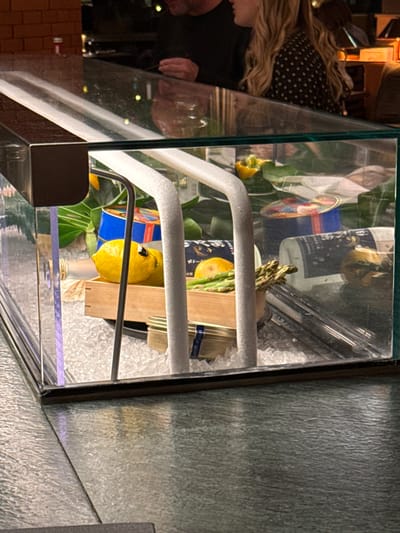 This image captures an indoor scene, likely within a restaurant or bar in Bordeaux, France. A clear, rectangular glass display case occupies the foreground, resting on a textured dark green or grey countertop. Inside the display, various food items are arranged on a bed of crushed ice. These items include at least two bright yellow lemons, one of which has googly eyes affixed, resembling a face. Also present are a bundle of green asparagus spears, several blue cylindrical cans with colorful labels, two bottles (one clear, one darker), small glass jars or tins (one with a blue-rimmed label reading "Gourmet de L'Atlantique"), and a light-colored wooden crate. Green foliage, possibly large leaves, is visible behind these items within the case. Two white, U-shaped support bars are positioned vertically inside the display. In the blurred background, behind the display case, two individuals are partially visible. To the left, a man wearing a dark top is seen from the chest up. To the right, a woman with long blonde hair, wearing a black top with white polka dots, is visible in profile. Their faces are indistinct, but their proximity suggests an interaction at a bar or counter. The lighting is dim, creating reflections on the glass surfaces.