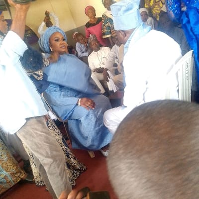 This image depicts an indoor social gathering in Ozuri, Nigeria. In the immediate foreground on the right, the top of a dark-skinned head is visible, partially obscuring the scene. To the left, a man in a light blue shirt and tan trousers stands with his left arm raised, holding an object.
The central figures are a woman and a man, both seated. The woman, positioned prominently to the left of the center, wears a light blue traditional dress and a matching gele (headwrap). She has visible makeup and a gold-colored bracelet on her left wrist. Her feet are visible, clad in shiny silver-colored shoes. She is seated on a light-colored chair, looking slightly to her right. The man, seated to her right with his back mostly to the viewer, wears a light blue traditional cap and a white traditional tunic and trousers. He is seated on a white plastic chair.
In the background, several individuals are seated and standing. These include men in white traditional attire, some with small caps, and women in various patterned and solid-colored traditional dresses (including pink). Children are also visible among the seated crowd. One man in the background, wearing white and a dark cap, appears to be holding a dark rectangular object. The environment features light-colored walls and a red-brown floor covering. A blue fabric is partially visible on the far right.