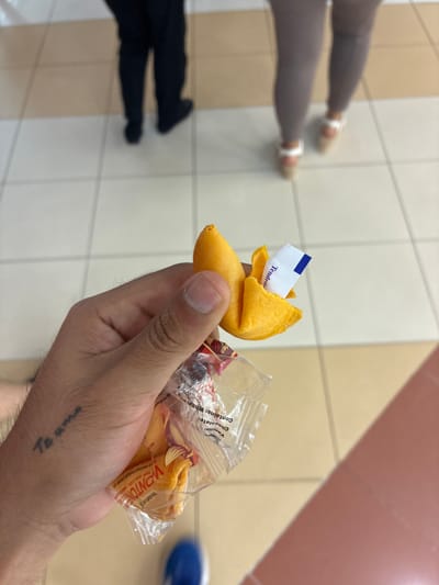 A person holds a fortune cookie in their hand, the wrapper containing another cookie. The fortune sticking out of the cookie reads "Tendrás". The tattoo "Te amo" is visible on the hand. In the background, there are two other people, one wearing black pants and the other wearing grey pants and sandals. The floor is tiled, and the background is blurry. The location cannot be determined from this image, but the fortune is in Spanish, suggesting a Spanish-speaking location. There is nothing in the image that indicates it is in the city of Coatzacoalcos, Mexico.
