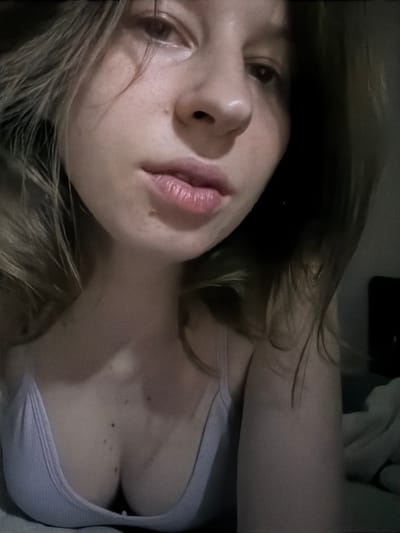 A close-up, low-angle, indoor portrait of a young woman is shown. She has light brown hair and is wearing a light purple tank top. The background is dark and blurred, with a hint of bedding visible on the right side. The image appears to have been taken in a dimly lit room. There is no discernible connection to Dongducheon-si, South Korea.