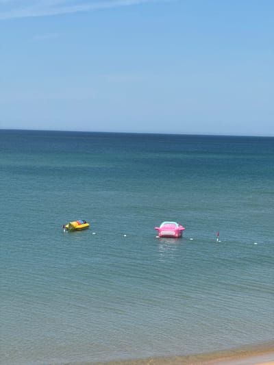 An aerial, wide shot captures a serene seascape with two inflatable water toys floating in the shallow, calm, blue-green water. The scene is oriented vertically, with the sky dominating the left side of the frame and the ocean extending to the right. The sky is a pale, hazy blue, suggesting a clear day with some atmospheric diffusion, possibly early morning or late afternoon given the soft light.

On the right side of the ocean, a sandy shoreline is visible, running diagonally from the top right corner of the image down towards the center. The sand appears light brown and is slightly wet near the water's edge, indicating a receding tide or gentle waves.

The first inflatable object, positioned closer to the foreground, is a bright pink, car-shaped toy. It has a distinct retro design with rounded edges and visible wheels. The second inflatable, located further out in the water, is a yellow, torpedo-shaped toy with red and blue stripes. It also has a small red and white flag attached.

Several small white buoys are dotted across the water, suggesting a designated area or a marker for a water activity. A single red flag on a pole is also visible near the buoys and the pink car. The water itself is mostly smooth, with faint ripples indicating minimal wind. There is no visible text in the image. The overall impression is one of a peaceful, recreational setting, likely a beach or a calm bay.