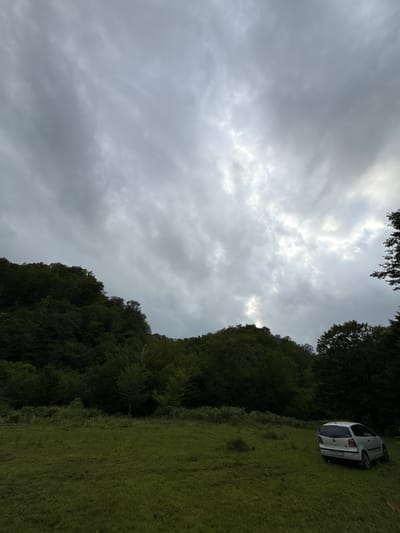 The image depicts a car parked on a grassy slope, adjacent to a dense forest. The sky is overcast with thick, gray clouds, suggesting a cloudy or potentially rainy day. The grass is a vibrant green, indicating a natural, outdoor setting. The car, a white hatchback, is positioned on the incline, with its wheels slightly sunk into the grass. A license plate is visible, but the text is too blurred to read.

The scene is devoid of people, emphasizing the solitary presence of the car within the natural landscape. The dense foliage of the forest creates a dark contrast against the brighter, overcast sky. The composition, with the car tilted and the sky dominating the upper portion of the image, evokes a sense of stillness and perhaps a touch of melancholy due to the weather. The time of day is difficult to ascertain precisely due to the cloud cover, but the overall lighting suggests it is daytime. The location cues, such as the lush greenery and dense forest, are consistent with a natural, potentially rural or wilderness area.
