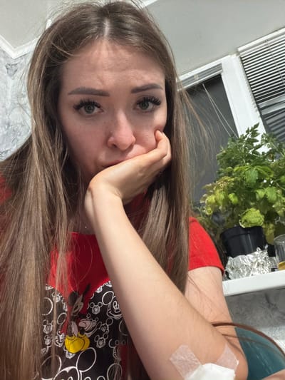 A young woman with long, brown hair and a red t-shirt featuring a Mickey Mouse pattern rests her head on her hand, looking directly at the camera with a somber expression. A bandage is visible on her forearm, held in place by medical tape. The background shows a window with blinds, a plant in a pot, and what appears to be a windowsill. The setting is indoors. No specific location in the city of Япрыково, Russia can be determined from the image.