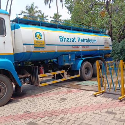 The image captures a Bharat Petroleum tanker truck parked on a paved surface with a herringbone pattern. The truck is predominantly white and blue, with the Bharat Petroleum logo prominently displayed on its side. The text "FLAMMABLE MOTOR SPIRIT" is also visible. An emergency technical panel is displayed on the tanker, providing information such as the phone number of the Marna Fire Department.

A yellow and blue safety barrier stands near the truck. The surrounding environment includes palm trees and other greenery. The truck appears to be at a fuel distribution facility, and the scene suggests an everyday operation related to fuel supply and transportation.