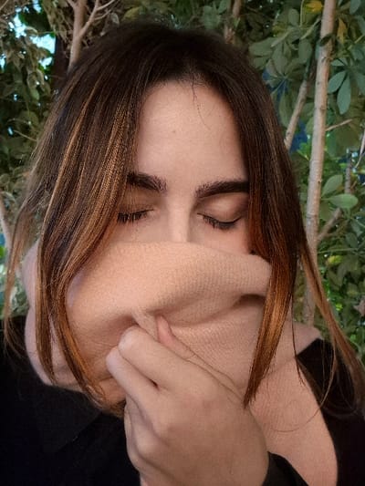 A woman with closed eyes and brown hair pulls at her cheek. Her skin is light-toned. A dark garment is partially visible. The background is an out-of-focus display of green foliage and woody branches. The image is oriented sideways, with the subject's face tilted towards the right.