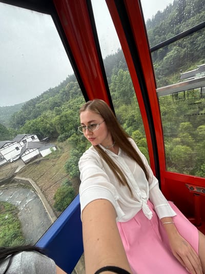 A person wearing glasses and a white blouse and a pink skirt is in an orange cable car. It appears to be raining outside, as indicated by the water droplets on the glass and the gray sky. Outside the cable car are lush green trees and a partially-visible white building with a dark roof. Based on the prompt information, the location is Yongding District, China.