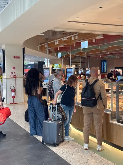 The image depicts an airport scene, likely Fiumicino Airport in Fiumicino, Italy, given the setting and context. Several people stand in line, most appearing to be travelers with luggage. A man with a shaved head wears a tan jacket and black backpack and looks towards the front of the line. A woman in a black shirt and jeans stands next to him. Behind them, another woman wearing a patterned shirt pulls a suitcase. A woman with curly hair stands at the end of the line in blue pants and a brown top. In the background are illuminated screens showing flight information and displays for restaurants. A fire extinguisher stands on the wall near the end of the line. The floor is gray tile, and the ceiling has a curved, modern design.