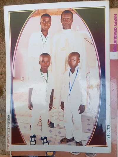 A group photo shows four males. From left to right, the first is a young man with a green lanyard, wearing a white collared shirt and white pants. The second is a young boy wearing a white shirt, white pants, and a green lanyard. The third is an adult male wearing a white priestly vestment with a cross-like design on it. The fourth is a young boy wearing a white shirt, white pants, and a blue lanyard. The background appears to be an indoor setting with light-colored walls and a tiled floor. Text on the right side of the photo reads "HAPPY GRADUATION" vertically. Text on the bottom left reads "GOD-DAY PIX .08134684511". Text on the bottom right reads "1/4/2018". The image is a photograph of a printed photograph. The location is stated to be Dutsen Alhaji, Nigeria.
