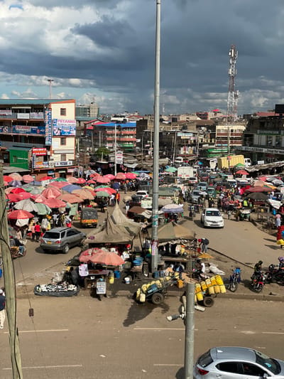 The scene takes place in Githurai, Kenya, at the intersection of two roads, near the coordinates -1.203 latitude and 36.918 longitude. The image shows a bustling outdoor market with numerous vendors selling their wares. The scene is dominated by a variety of stalls, some of which are sheltered by temporary canopies. There are numerous brightly colored umbrellas scattered throughout the market. Hundreds of people are in the market, some are vendors, others are customers, and some are simply walking through. Cars are parked in a haphazard manner on both sides of the road. In the background, there are buildings, including a tall building with the name "PETANNS TECHNICAL COLLEGE" in large lettering. The weather appears partly cloudy with a hint of rain. There is a cell phone tower visible in the background. The street itself is paved and there is a median strip in the middle, separating the two lanes of traffic.