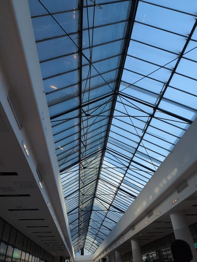 The image depicts the interior of the Jumbo shopping center in Vantaa, Helsinki, Finland. The photograph is taken from a low angle, looking up at the expansive glass ceiling and the architectural structure supporting it. The ceiling is composed of numerous rectangular glass panels framed by a dark, angular metal framework. The sky is visible through the glass, appearing as a muted blue, suggesting it is either early morning, late evening, or overcast. Lights are visible through the glass panels, indicating interior lighting within the shopping center.

On the left and right sides of the image, parts of the shopping center's interior architecture are visible. These include white, curved structural elements that appear to be part of the building's upper levels or a mezzanine. Small, rectangular lights are embedded along these white structures. There are also ventilation grates visible on these surfaces. Below the glass ceiling, on the far left and right, are glimpses of the shopping center's interior, with dark storefronts or displays visible. A dark silhouette of a person's head and shoulders is visible in the lower right corner of the frame, partially obscuring the view of the interior. The overall impression is one of a modern, spacious, and well-lit commercial environment.