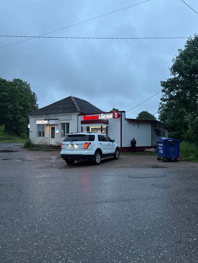 A white Ford Explorer SUV is parked in front of a small, light-colored building with a dark roof, likely a storefront, in Valday, Russia. The building has a sign that reads "Ozon" in neon lights on the left side and another sign with red lettering on the right side that appears to say "Коктейль Белое". A blue dumpster is located to the right of the building, and a person in dark clothing is standing near the rear entrance of the building, facing away from the camera. The ground is wet and appears to be asphalt, with puddles reflecting the dim, overcast sky. Trees are visible in the background on the left and right sides of the image. The overall scene suggests a commercial area on a cloudy day.