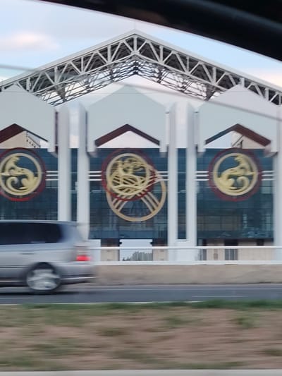 The media file depicts a building with a distinctive architectural design, featuring a large, white, triangular roof structure supported by a metal framework. The facade of the building is a combination of white panels and dark blue glass. Prominently displayed on the facade are circular emblems in gold and red, each with a stylized depiction of a creature, possibly a mythological being or animal. To the left of the building, a silver car is blurred due to motion, indicating that the image was taken from a moving vehicle. The car is on a road in the foreground, with a grassy area beyond the road. The overall scene suggests a daytime setting, possibly late afternoon, given the lighting. The sky appears to be a pale blue with some clouds. There is no visible text in the image. The location context of Бактуу-Долоноту, Kyrgyzstan, might relate to the architectural style or the emblems featured on the building.