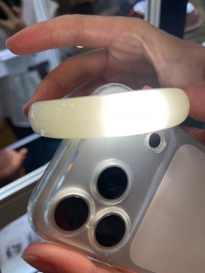 A close-up shot shows a hand holding a white, translucent bangle over the camera lens of a clear phone case. The bangle is illuminated from behind by a bright light source, making it appear almost opaque in the center and translucent at the edges. The phone case reveals the phone's camera lenses and a textured backing. The background is blurred, but suggests an indoor setting with display cases and other objects that are out of focus. The lighting and composition suggest that the bangle is being examined or showcased.