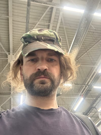 A man with light brown, wavy hair, a beard, and a mustache wears a camouflage baseball cap and a dark purple t-shirt. He is looking directly at the camera. The background consists of a ceiling with exposed metal beams and fluorescent lights, typical of an indoor public space. The image appears to have been taken from a low angle, looking upwards. The location is unknown, but is not Biysk, Russia.