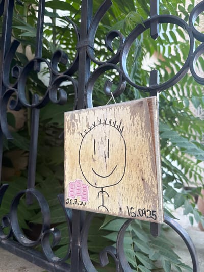 A close-up, eye-level outdoor shot captures a wooden sign hanging on a black metal gate. The sign, weathered and with peeling paint, displays a child-like drawing of a smiley face with a simple stick figure body. Below the drawing, two dates are handwritten: "26.7.10" on the left, accompanied by a pink sticker with three "OH" shapes, and "16.09.25" on the right. Lush green foliage is visible behind the gate and sign, suggesting a garden or courtyard setting. The ornate scrollwork of the gate is a characteristic feature of traditional architecture found in the historic city center of Nicosia, Cyprus.