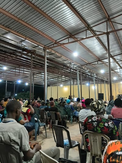 A gathering of people sit in rows of chairs under a large metal roof supported by tall beams. Overhead lighting illuminates the space. Individuals are dressed in casual clothing, and some wear patterned head coverings. The scene suggests a community event or meeting in a simple, covered area.