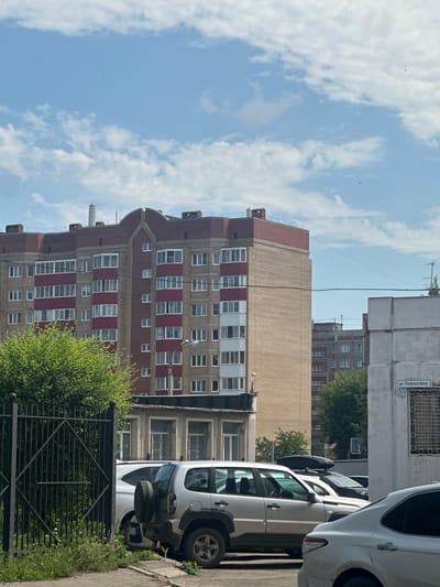 A daylight, outdoor, eye-level medium shot shows a residential street scene in Magnitogorsk, Russia. The main subject is a multi-story brick apartment building with red trim on its balconies and roof. In the foreground, a black metal fence with diamond-shaped patterns encloses lush green foliage and grass. Beyond the fence, several cars are parked on a paved area. A silver SUV with a spare tire mounted on its rear door is prominent, alongside other vehicles partially visible. To the right, a white building displays a street sign reading "ул. Тевосьяна 25/1." The sky above is blue with scattered white clouds.