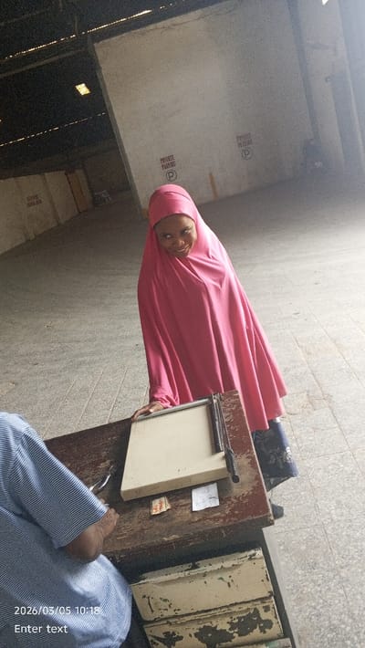The scene is set in an indoor or semi-enclosed commercial space in Dakata, Nigeria, likely a print shop or similar establishment, characterized by concrete walls and a high corrugated roof structure partially visible at the top.
In the foreground, a person is seated to the left, with only their right shoulder and arm visible. This individual wears a light blue and white striped long-sleeved shirt and holds a small metallic tool, resembling pliers or a staple remover, in their right hand. They are positioned at a well-worn, dark brown wooden table with visibly chipped and faded paint, revealing lighter wood beneath. The table features light-colored drawers with similar paint wear.
On the table, an off-white manual paper cutter with a black handle is centrally placed. To the left of the paper cutter, a small stack of what appears to be folded papers or currency is visible, with faint red markings on one piece. A small, crumpled piece of white paper with illegible writing lies next to it.
Standing behind the table on the right side of the frame is a woman, smiling directly towards the camera. She wears a vibrant pink full-body abaya or hijab that covers her head and body, and dark blue trousers or a skirt with a subtle floral pattern is visible underneath the abaya. Her right hand rests on the table near the paper cutter.
The background consists of light grey concrete walls with repetitive red stenciled text, "PRIVATE PARKING," accompanied by a circular parking symbol. The floor is light-colored and paved, possibly with tiles or concrete slabs. The image includes a digital timestamp "2026/03/05 10:18" in the bottom left corner.