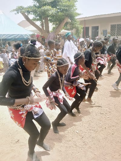 An outdoor cultural performance is captured during daylight hours in Masvingo, Zimbabwe. In the foreground, a line of approximately eight individuals, primarily young girls and adolescents, perform a coordinated dance on a dry, light brown earth surface. They are dressed in black long-sleeved tops, black leggings, and patterned fabric wraps in red, white, and black tied around their waists. Accessories include multiple strands of light-colored bead necklaces, bracelets, and headbands. Their posture involves bent knees and hands raised with palms partially visible, indicative of rhythmic movement. Footwear varies from dark soft shoes to apparent bare feet.

In the mid-ground, a male adult in a white flowing garment and a blue-green cap stands, observing the performers. Several younger children in colorful patterned clothing are also present. The background shows numerous spectators, some standing and others seated under a light blue tent canopy on the left. A large, mature tree with sparse green foliage occupies the center-background. To the right, a white single-story building with multiple windows and a gray roof is visible, featuring a colorful graphic design on its wall. The sky is bright and clear.