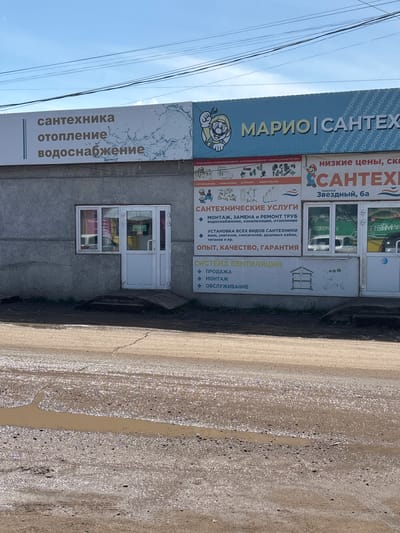 Here is a description of the image:
The image shows the storefront of a plumbing and heating supply business in Ust-Ordynsky, Russia. The business is called "Mario Santex" and offers services such as installation, repair, and sales of plumbing, heating, and ventilation systems. The building is a gray, one-story structure with a white door and windows. The street in front of the building is unpaved and muddy.