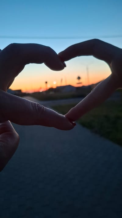 Two hands are silhouetted against a twilight sky, forming a heart shape. The background reveals a blurred landscape with a path, buildings, and distant lights, suggesting an urban or suburban setting. The sky displays a gradient of blue to orange hues, indicative of sunset or sunrise. There is no discernible location in the city of Stowbtsy, Belarus within the image.