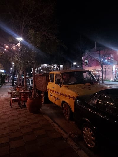 This nighttime image captures a lively street scene, likely in an urban area. The focal point is a vintage-style yellow car, possibly a replica or a classic model, parked on a paved sidewalk. The car features a distinctive red patterned decal along its side and clearly displays the black text "Sheshi Skënderbej". Part of a dark-colored car is visible behind it.
The street is lined with bare trees, their branches silhouetted against the dark sky. Numerous streetlights illuminate the scene, casting bright beams and creating lens flares. Further down the street, lights from buildings and other establishments indicate a bustling commercial or residential area. On the sidewalk near the yellow car, outdoor seating, including tables and chairs, suggests a cafe or restaurant. A large, reddish-brown pot or planter sits on the brick-paved ground.
The time of day is clearly night, with all illumination coming from artificial light sources. No individuals are distinctly visible, but the setup implies an active area. The visible text "Sheshi Skënderbej" serves as a strong location cue, referring to Skanderbeg Square in Tiranë, Albania, aligning with the provided context.