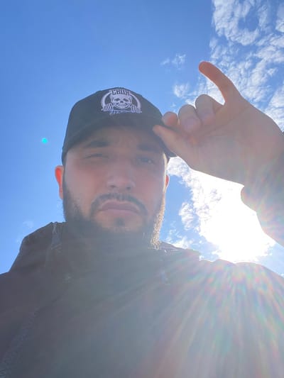 A low-angle, selfie-style photograph shows a man with medium skin tone, a dark beard, and a black baseball cap with a white skull and barbell logo. He is partially backlit by the sun, causing lens flare. He is pointing with his left index finger towards the blue sky with scattered white clouds. The man is wearing a dark jacket. The location is Koskolovo, Russia.