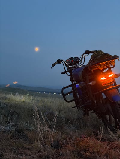A motorcycle is parked in a grassy, hilly landscape at dusk. The sky is a deep blue with a faint, hazy glow, possibly from distant lights or a celestial body. The motorcycle's taillights are illuminated with a bright orange glow, and its headlights are also on, casting a light on the dry grass in front of it. In the distance, rolling hills are silhouetted against the twilight sky, with faint lights suggesting a settlement or scattered dwellings. The overall scene evokes a sense of quiet isolation and a journey potentially ending or paused in a remote area.