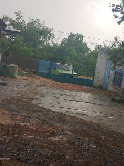 In Pryvilne, Ukraine, a courtyard scene unfolds under an overcast sky. A weathered green and blue metal gate stands partially open, revealing a vintage green vehicle parked behind it. To the left of the gate, a cylindrical, green-painted well structure with a metal pump is visible. The ground in the foreground is a mixture of dirt, gravel, and what appears to be a wet, cracked concrete or asphalt surface. In the background, lush green trees form a dense natural backdrop, interspersed with parts of buildings. One building, on the right side of the frame, has a pale, textured wall with a blue-framed window. The overall impression is one of a rural or semi-rural setting, possibly a farmstead or an older residential area.