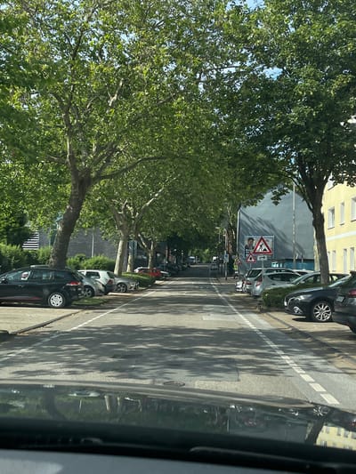 The image depicts a street in Essen, Germany, lined with trees and parked cars on both sides. The street is paved and has lane markings. On the left side of the street, several cars are parked along the curb, with a black station wagon in the foreground. On the right side, a row of cars is also parked, including silver and black vehicles. Beyond the parked cars, buildings are visible. One building is a pale yellow, and another is a darker gray. There are traffic signs present, including a warning sign with figures working and a triangular sign with a pedestrian and a bicycle symbol. The scene is illuminated by sunlight, casting shadows from the trees onto the road. The overall impression is of a residential or commercial street on a sunny day.