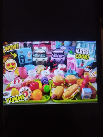 The image displays a collection of toy food items and kitchen appliances arranged on a bright green tray. The toy food includes hamburgers, a hot dog, a sandwich, donuts, fruits such as apples and grapes, and what appears to be a carton of milk. The kitchen appliances include a coffee maker, a blender, and a pink kettle. Text overlays on the image read "WOW!", "YUMMY", and "LET'S COOK!". The background shows additional toy kitchen items, primarily in pink and blue, suggesting a play kitchen setting. The image is a still from a video, and there is no indication of a specific location in Özgön, Kyrgyzstan.