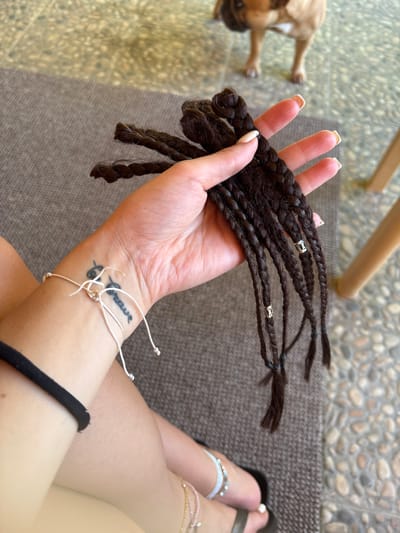 A close-up shot captures a person's hand holding a bunch of dark brown braided hair extensions. The braids are adorned with small, silver beads. The person's hand has manicured nails with a light color polish. A tattoo is visible on the person's wrist, and a thin white string bracelet is tied around it. In the background, a small, light-brown dog lies on a textured brown mat. The ground behind the dog is covered with a mosaic of small, multi-colored stones. A wooden chair leg is partially visible in the upper portion of the frame. The lighting suggests it is daytime, possibly outdoors given the stone flooring and mat. The scene appears relaxed and casual.