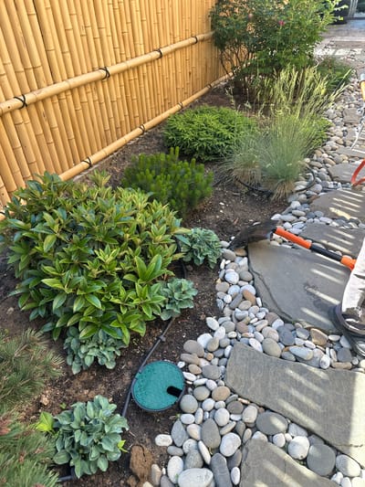 The image depicts an outdoor garden scene, likely in a residential setting, characterized by a bamboo fence that forms a backdrop. In the foreground, there is a variety of plants, including a lush green shrub with broad leaves, smaller evergreen plants, and some ornamental grasses. The ground is covered with a mix of soil, decorative pebbles, and flat stone pavers, creating a pathway or garden bed border. A green irrigation system access point is visible in the soil. To the right, a person's lower legs and feet are visible, wearing dark pants and shoes, and they are holding a shovel with an orange handle, suggesting active gardening work is in progress. The lighting suggests it is daytime, with some shadows cast by the plants and pavers. The overall impression is one of a well-maintained, yet actively tended, garden space.