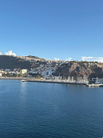 The image displays a panoramic view of San Sebastián De La Gomera, Spain, a coastal town nestled against arid hillsides overlooking a bay. The upper portion of the frame is dominated by a clear, bright blue sky with sparse white cumulus clouds. The town's buildings, primarily residential and multi-story, are densely constructed across the green-brown slopes, exhibiting a range of pastel colors including white, yellow, orange, and terracotta. A single white wind turbine is visible on a distant hilltop to the far left.
The lower half of the image features the deep blue, rippled waters of the bay. On the left side of the waterfront, a sandy beach is visible, with a line of white buoys delineating a section of the water. This beach is backed by a promenade lined with palm trees. Extending into the bay from the right, a dark-colored breakwater forms a marina. The marina is densely populated with numerous sailing yachts and recreational boats, characterized by their white hulls and numerous vertical masts. To the immediate right of the marina, additional port infrastructure, including larger dock structures and what appears to be a boat lift, is present. No individuals or specific actions are discernible within the frame.