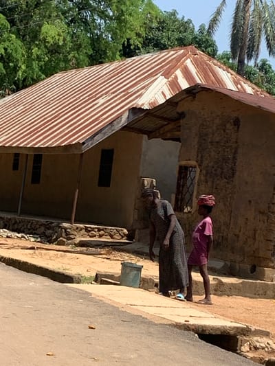 In Fada, Nigeria, an elderly woman in a floral dress stands on a dirt and stone-lined path beside a dilapidated building, while a young girl in pink stands beside her. The building has a corrugated metal roof and small windows. The environment includes trees, a road, and other vegetation.